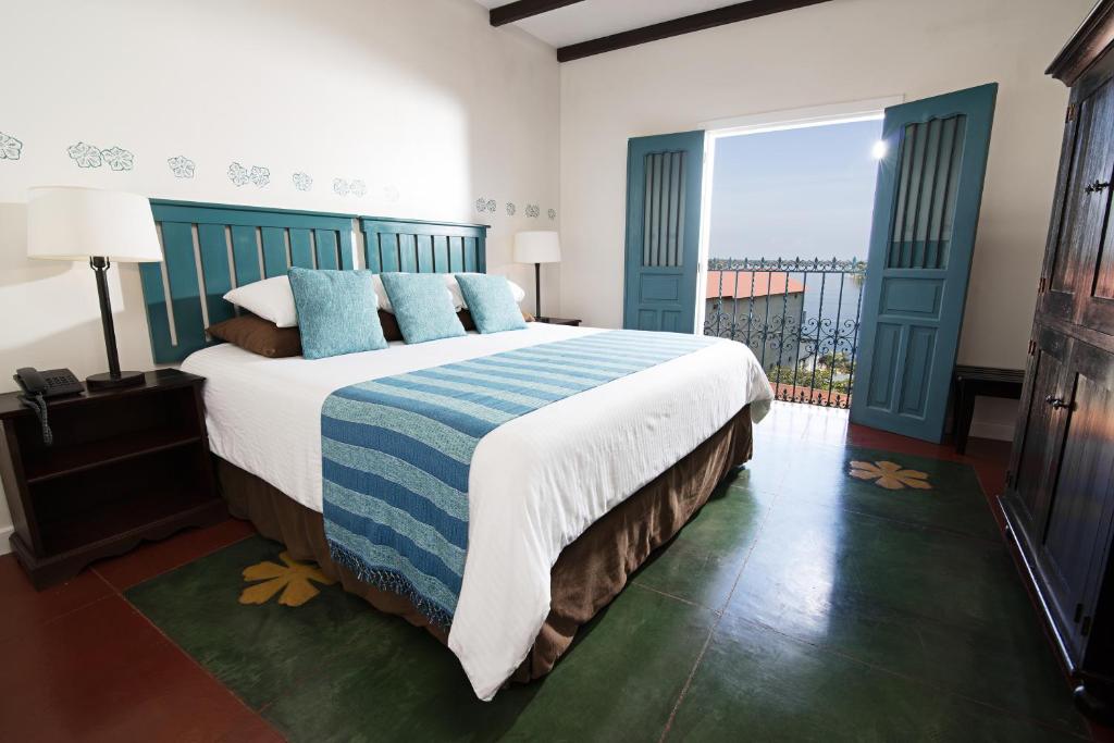 Hotel bedroom with teal headboard and blue striped throw; doors open to a balcony with an ocean view.