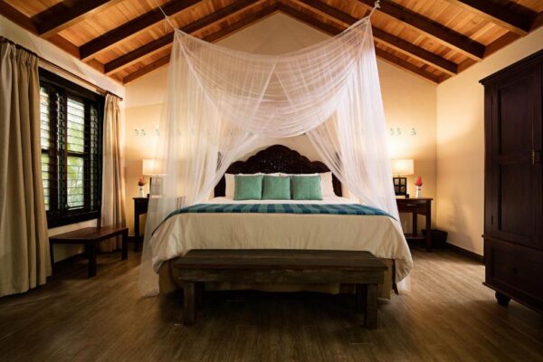 Bedroom with a four-poster bed draped in sheer white netting, teal throw pillows, and dark wooden furniture.