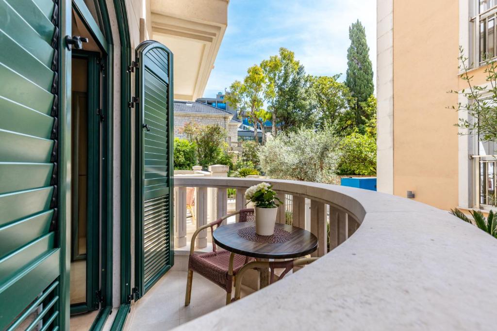 Balcony with a round wooden table and wicker chair, a potted white flower, and green shutters with a leafy courtyard beyond.