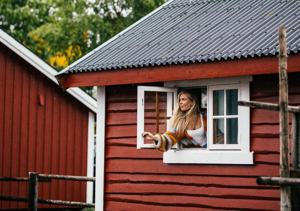 Woman leaning out of a white-framed window of a red wooden cottage, wearing a striped cardigan and white top on a sunny day.