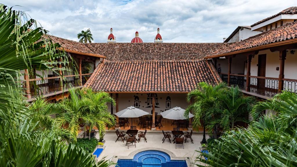 Two-story tropical hotel courtyard with clay-tiled roof, palm trees, and a small curved blue pool in the foreground; tables and umbrellas surround the area in the seating courtyard.