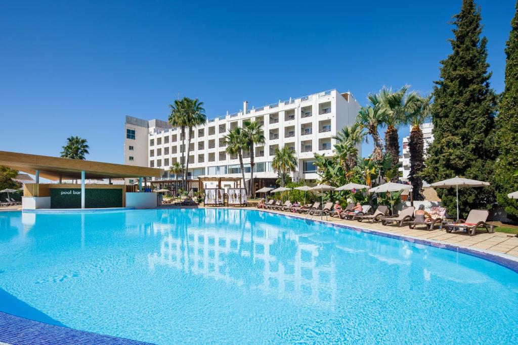 Hotel pool area on a sunny day: blue water, lounge chairs with umbrellas, and palm trees with a white multi-story building in the background.