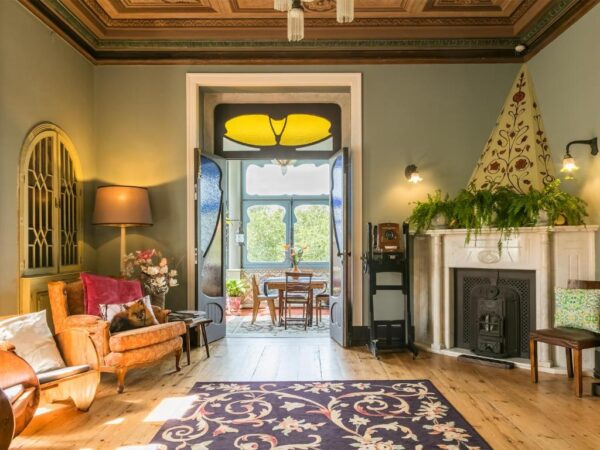 Sunny vintage living room with green walls, orange chairs, a cat on a cushion, and open double doors to a sunlit patio.