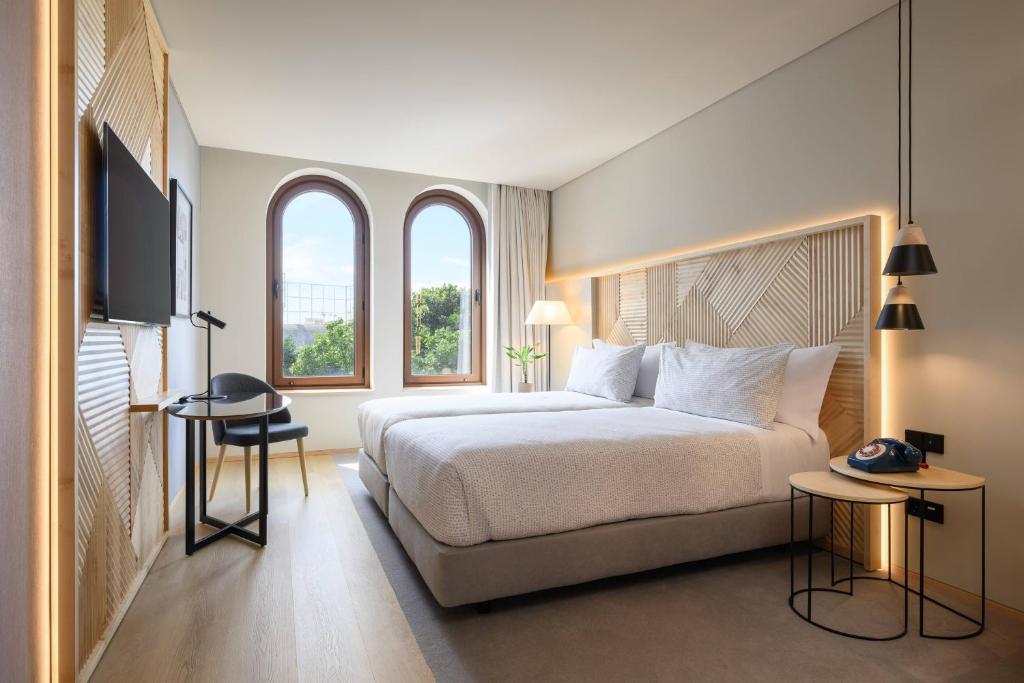Modern hotel room with a king bed, geometric headboard, soft lighting, and two arched windows overlooking greenery; includes a small desk and chair nearby.