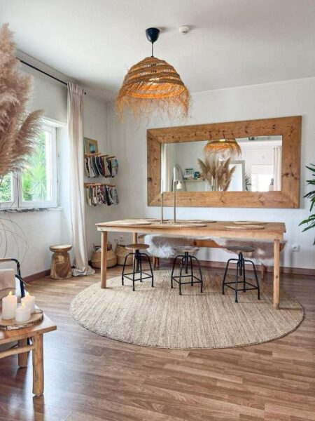 Boho-inspired dining/studio space with a long wooden table, black stools, and a circular jute rug, plus a large wooden-framed mirror and a woven pendant light above.