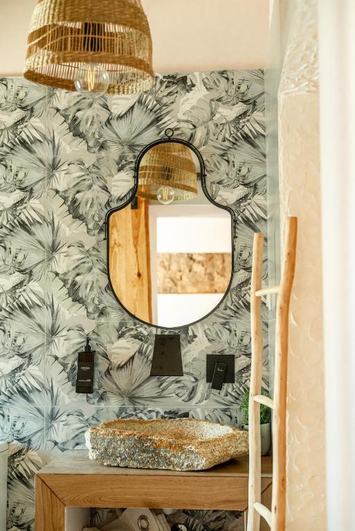Wood vanity with a stone sink and oval mirror, tropical palm wallpaper, and a woven pendant light above.