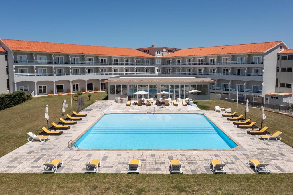Three-story hotel surrounding an outdoor rectangular pool with rows of yellow lounge chairs, white umbrellas, and a glass-walled central lobby on a sunny day.