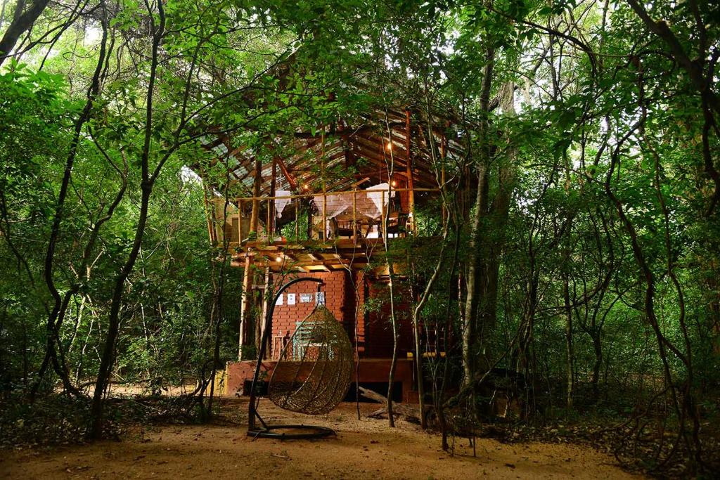 Two-story wooden treehouse perched among dense green trees, with a brick base and a hanging wicker chair in front on the ground.