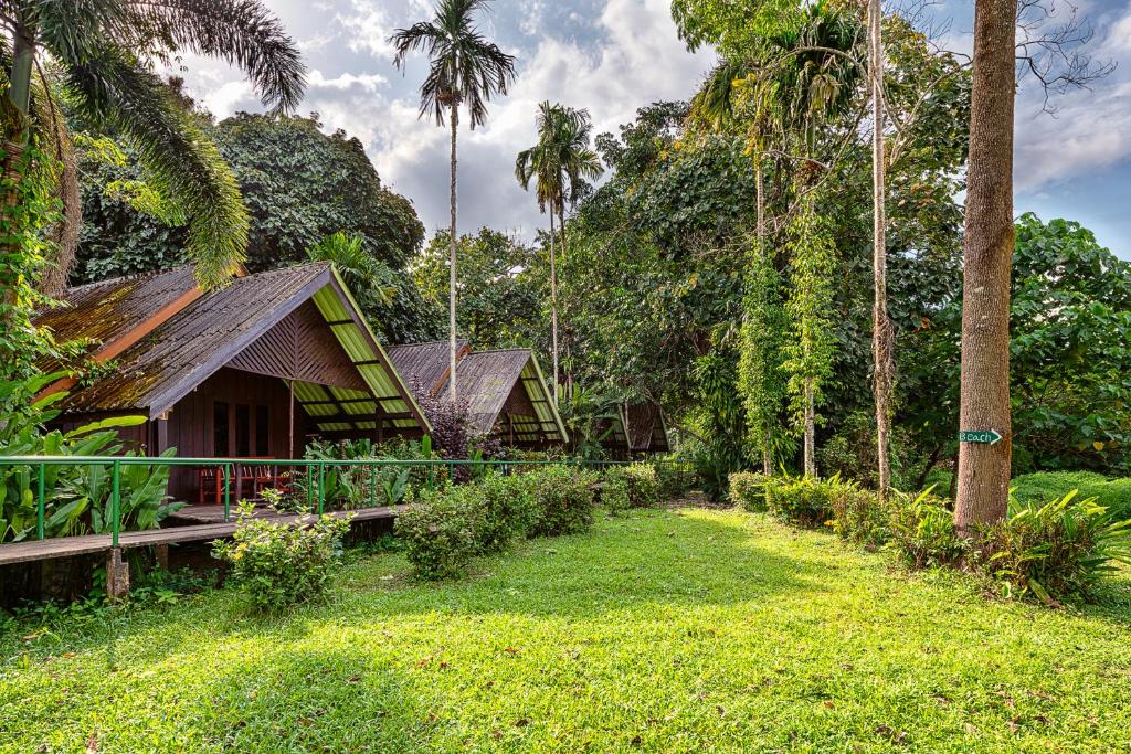 Tropical resort with wooden cabins and thatched roofs set among palm trees and green lawn. A path and railing lead to the cabins on a sunny day.
