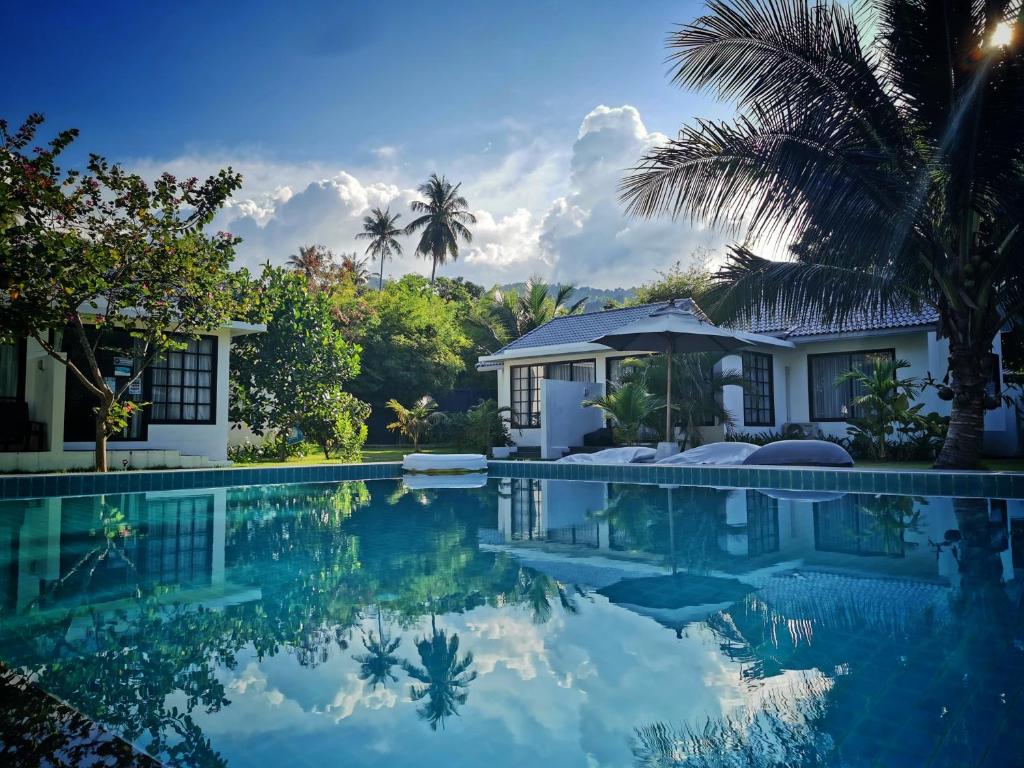 Resort pool surrounded by palm trees and white bungalow-style cabins, with a bright blue sky and clouds reflected in the water.