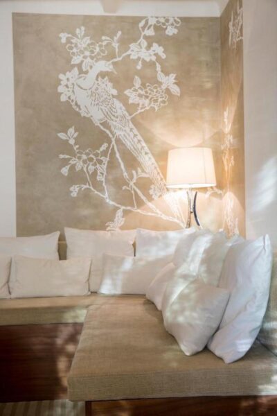 Corner seating area with a beige sectional and white cushions, against a wall featuring a white floral branch design and a wall-mounted lamp casting warm light.