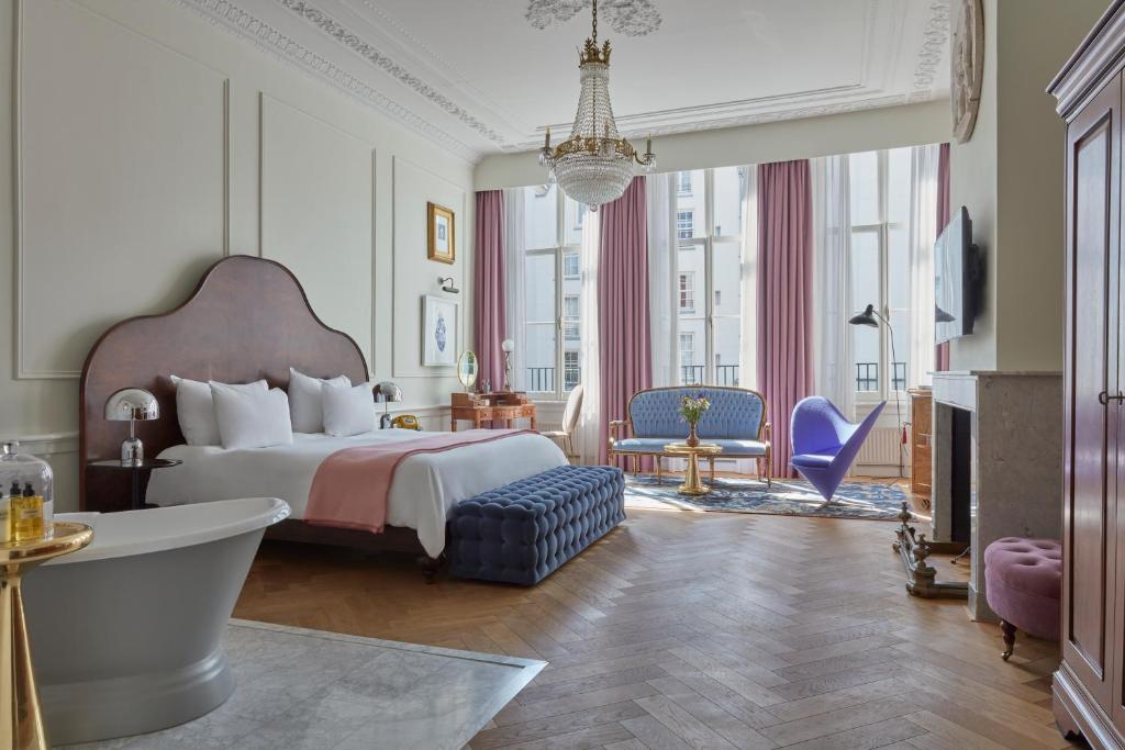 Luxurious hotel suite with a large bed, tufted navy bench, crystal chandelier, and seating area by tall windows with pink drapes.