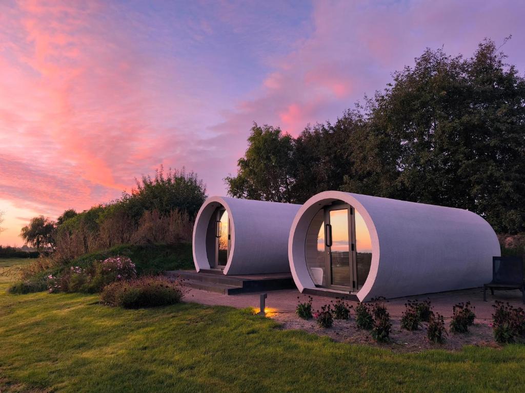 Two cylindrical pod houses with glass doors sit on a grassy landscape at sunset, pink and purple sky overhead.