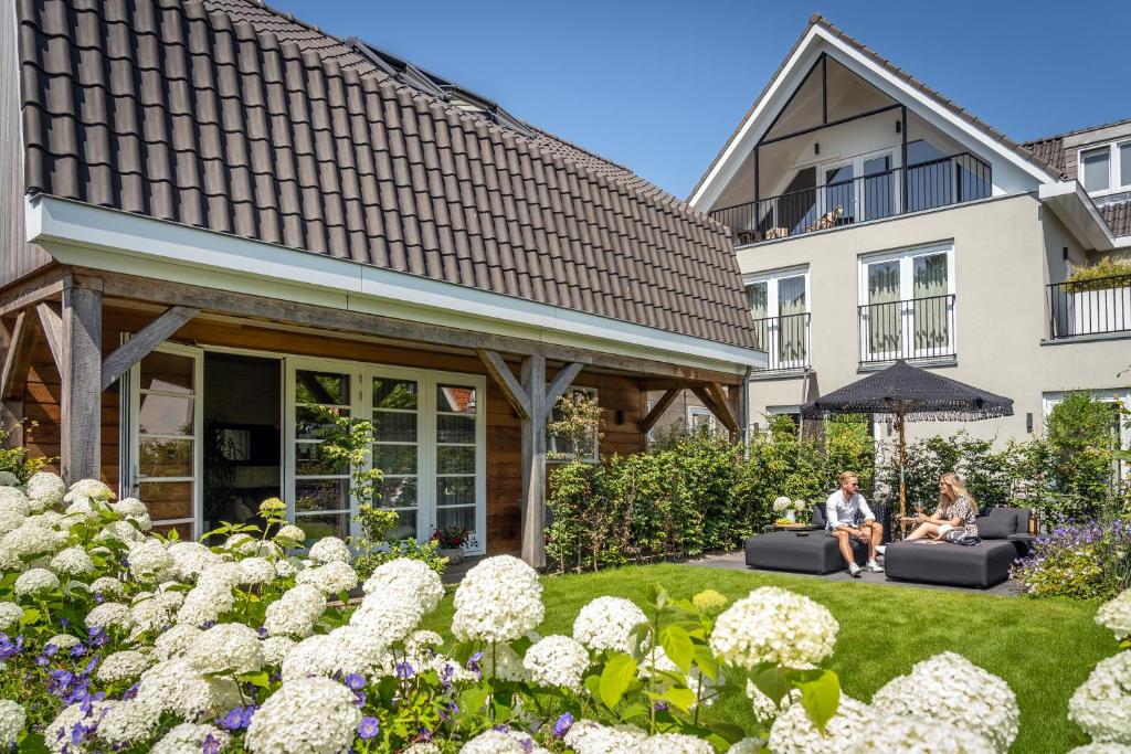 Sunny backyard with white hydrangeas in the foreground, a wooden patio extension, and two people relaxing on gray outdoor sofas under a parasol by the lawn.