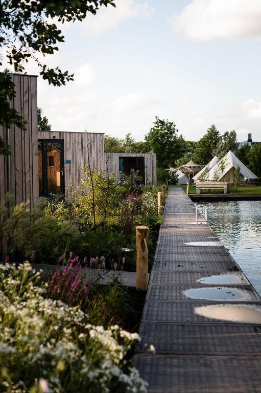Wooden boardwalk runs along a lake with small wooden cabins and white tents in the distance, surrounded by garden plants under a blue sky.