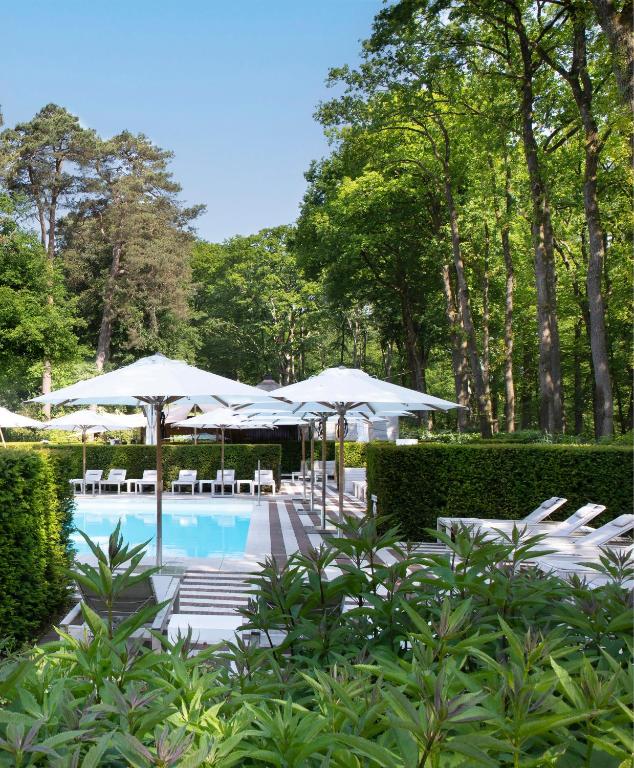 Outdoor pool area with white umbrellas and lounge chairs surrounded by manicured hedges and tall trees on a sunny day