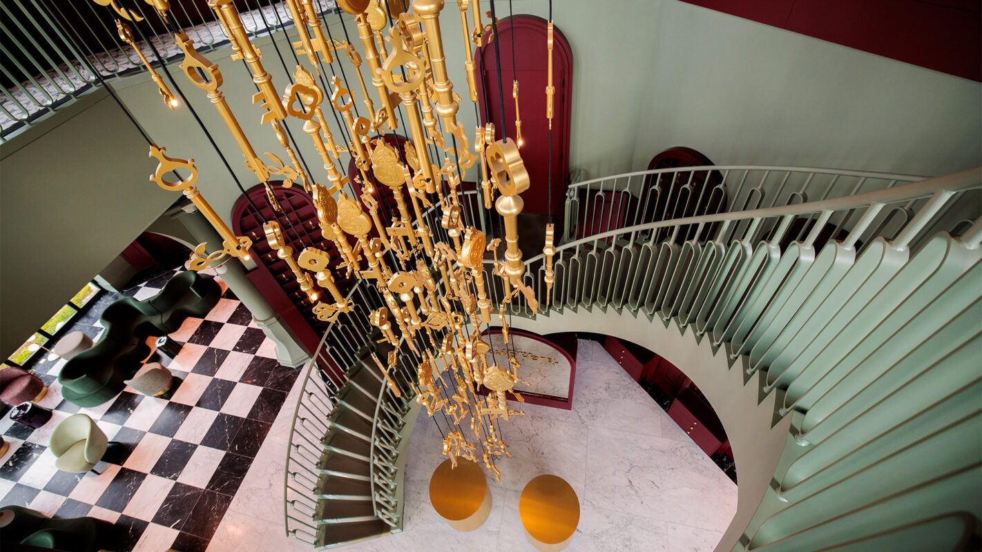 Wide-angle view of a spiraling staircase with a green railing and a dramatic gold chandelier hanging above, against a marble floor with black-and-white tiles and yellow circular accents in a stylish lobby.