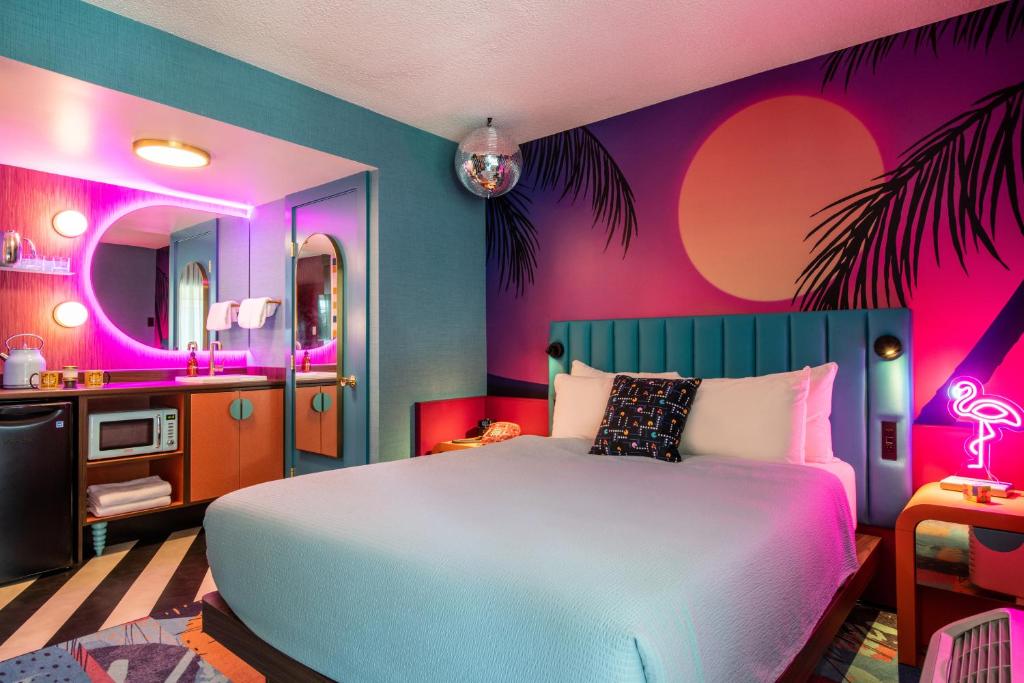 Colorful hotel room with a turquoise bed, neon pink lighting, and a tropical mural with palm silhouettes and a setting sun; kitchenette area nearby.