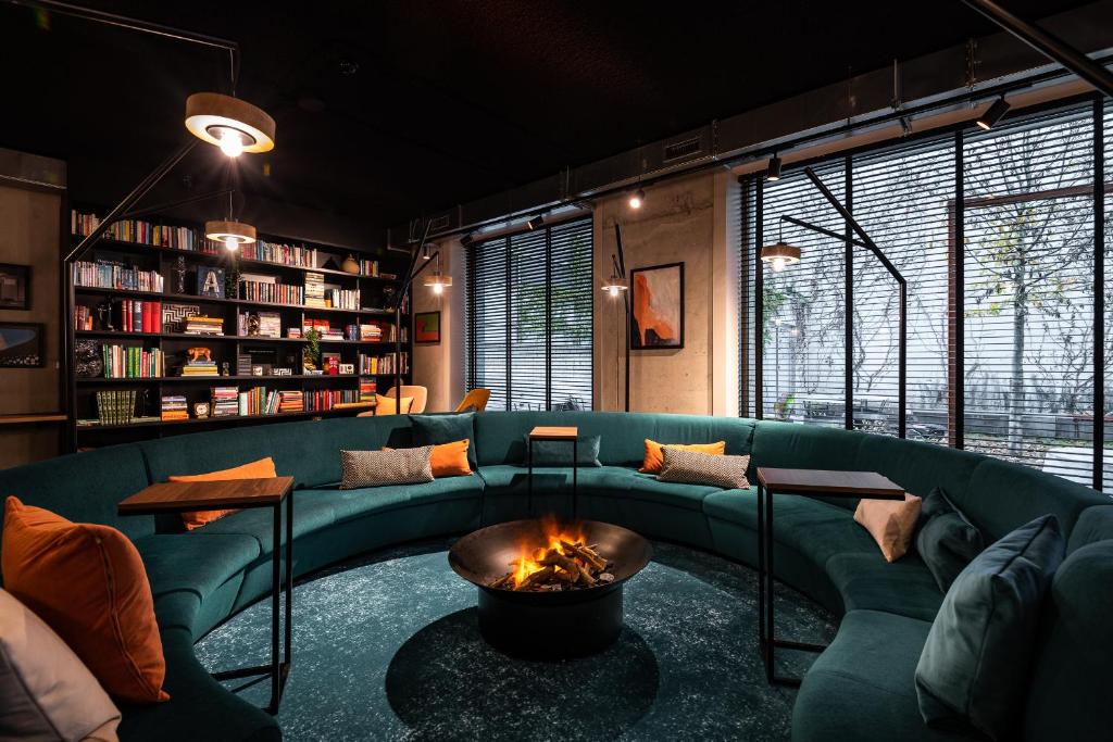 Circular teal sofa seating around a lit fire pit in a modern lounge with bookshelves and large windows.
