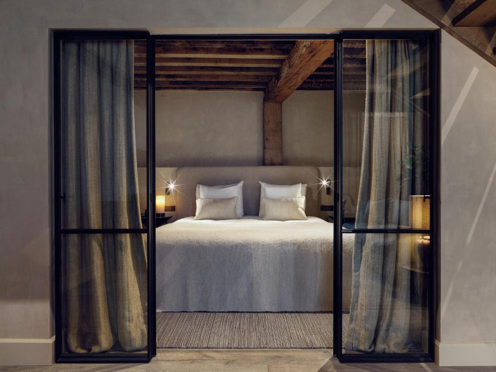 King-size bed with white bedding and beige pillows seen through dark-framed glass doors in a modern hotel room with rustic wooden beams above.