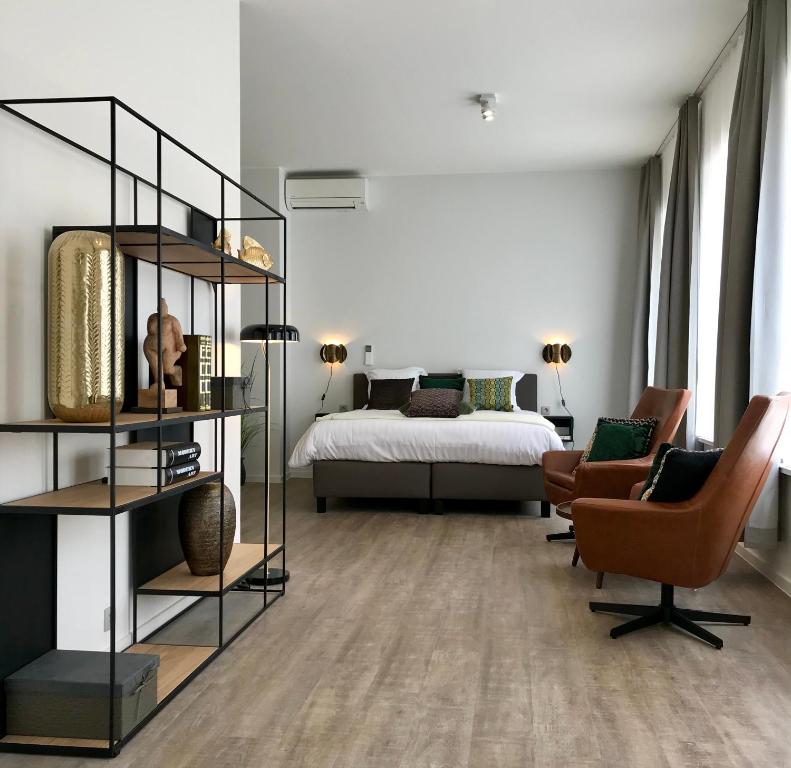 Modern hotel room with a king bed, two brown lounge chairs, and a metal open shelving unit on the left side of the room.