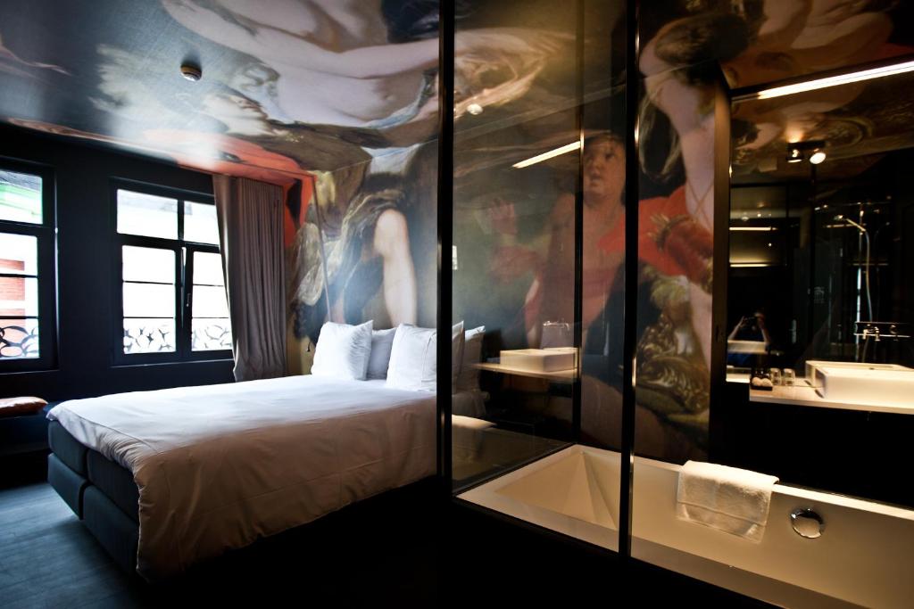 Hotel bedroom with a white duvet, pillows, and a glass-walled bathroom feature reflecting a colorful mural on the ceiling.