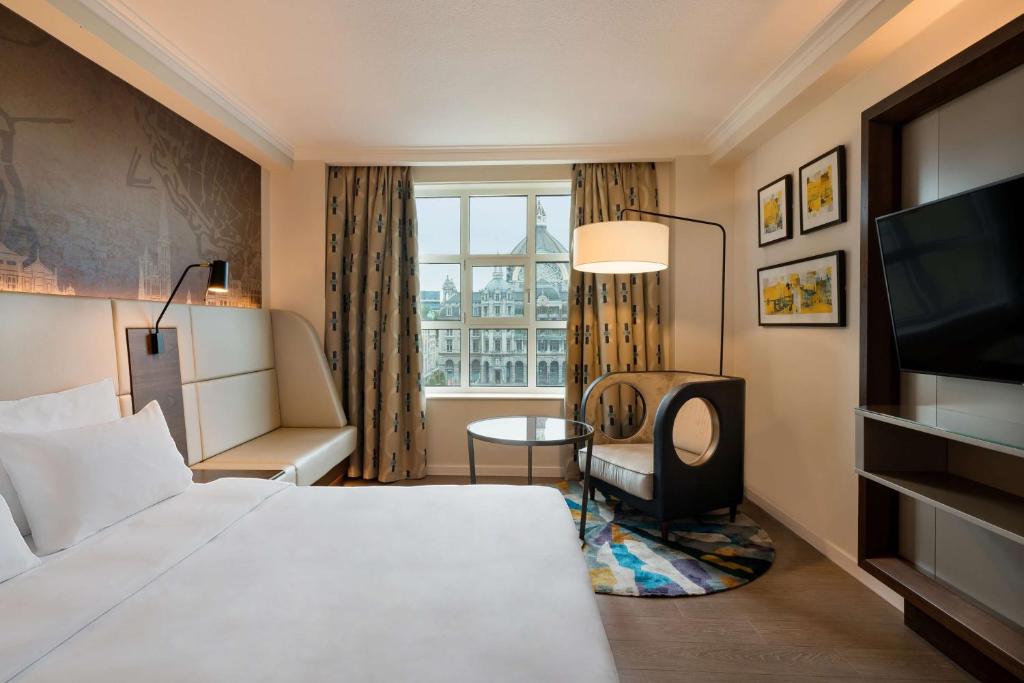 Hotel room with a white bed, seating nook by a window, and a round glass table under a curved floor lamp, city view outside the window.
