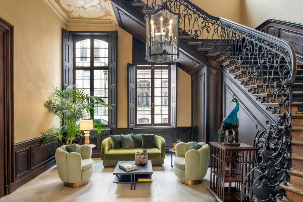 Luxurious living area with green sofa and rounded chairs, palm plant, and a wrought-iron staircase in a stately room.
