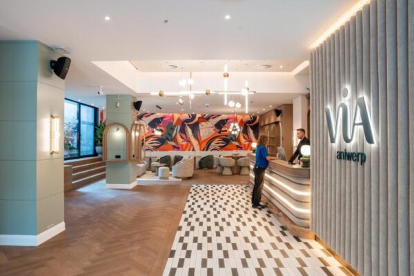 Reception area of a modern hotel lobby: a staff member assists a guest at the curved reception counter, with a colorful mural behind.