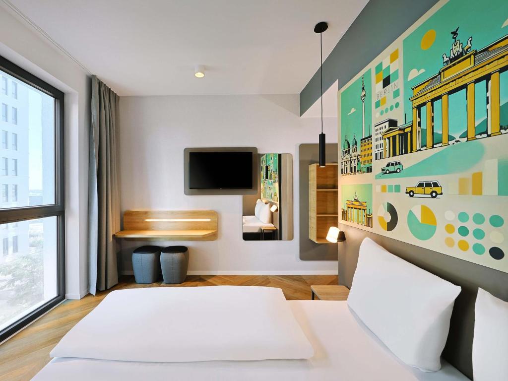 Hotel room with a large colorful mural of Berlin landmarks above the bed, white linens, and a window with gray curtains on the left.