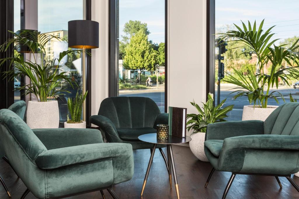 Cozy modern waiting area with green velvet chairs, a small round table, and potted plants by large windows.