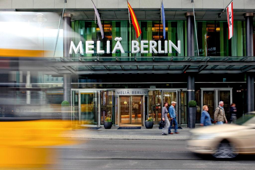 Meliá Berlin hotel entrance with glass doors and a storefront sign, pedestrians on the sidewalk, and blurred yellow taxis passing by.