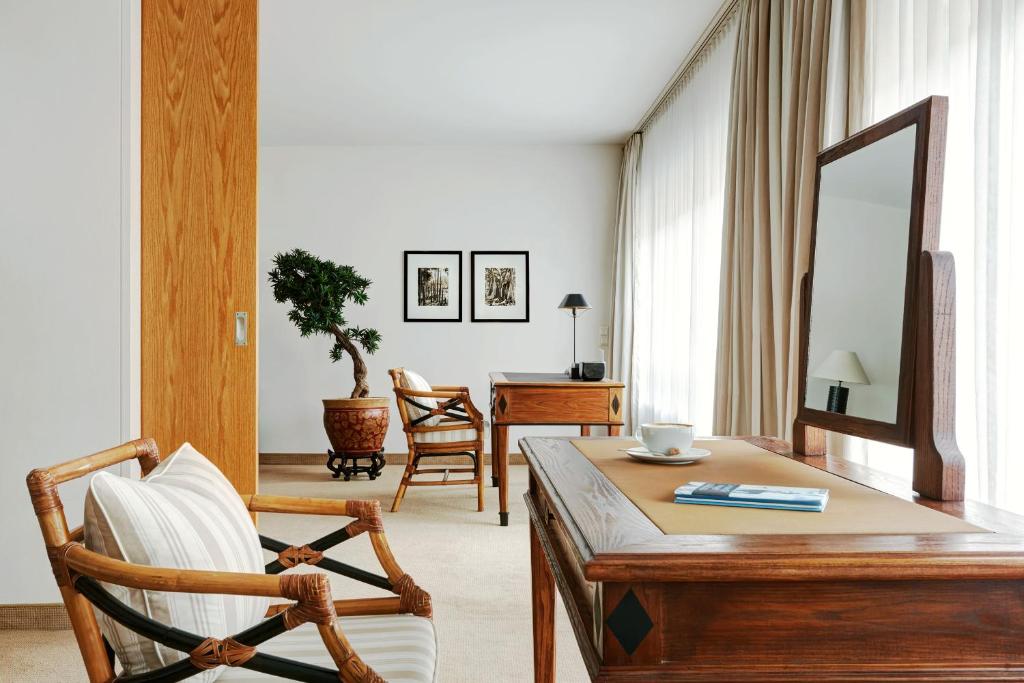 Bright hotel-style living area with a wooden desk, large mirror, and wicker chairs beside a potted plant in natural light.