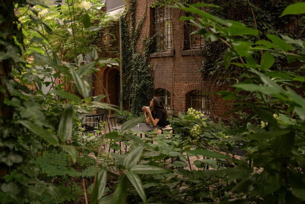 Person sitting at an outdoor table in a narrow garden courtyard, surrounded by greenery and ivy-covered brick walls.
