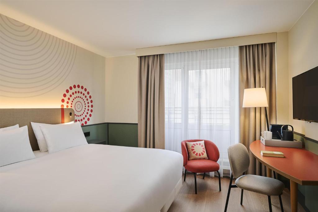 Hotel room featuring a king bed, red accent chair, desk, and a large window with sheer curtains.