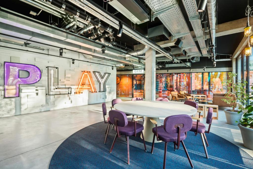 Open modern coworking area with a round white table and purple chairs, a neon 'PLAY' sign on a white brick wall, and an industrial ceiling.