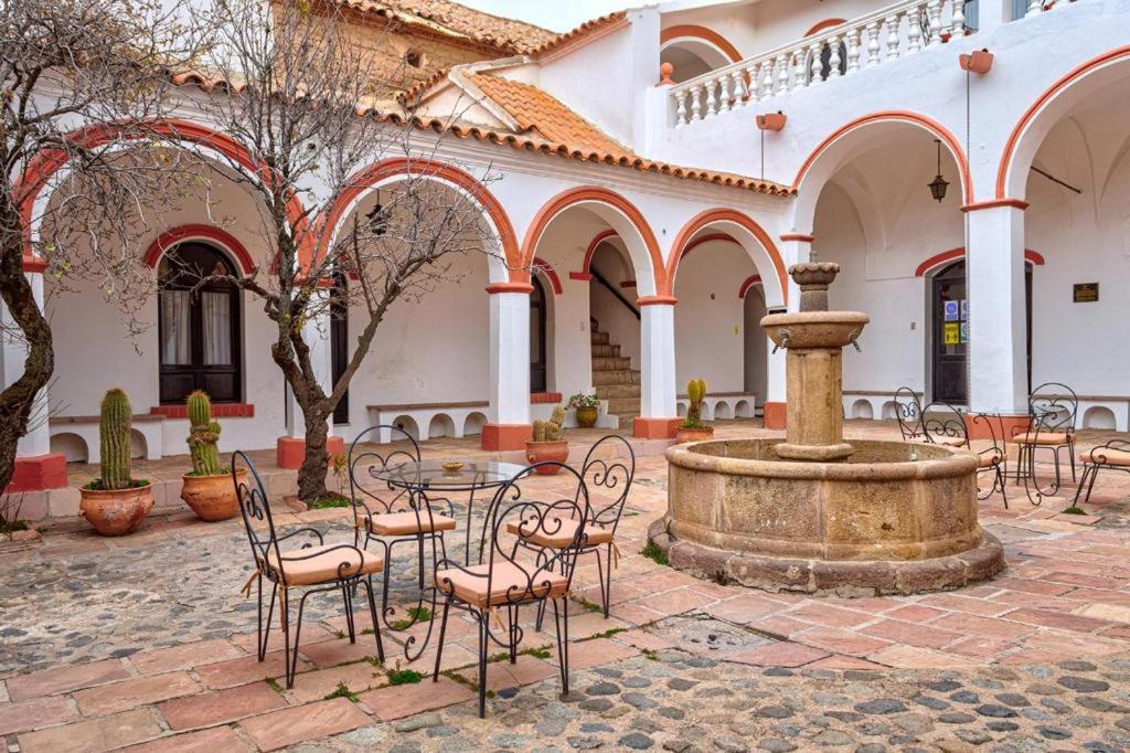 Arched white courtyard with red trim, a central stone fountain, and metal chairs around potted cacti.