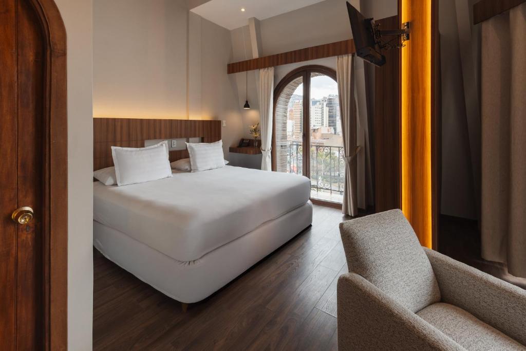 Hotel room with a king-size bed, armchair, and a balcony with a city view through arched doors on the right.