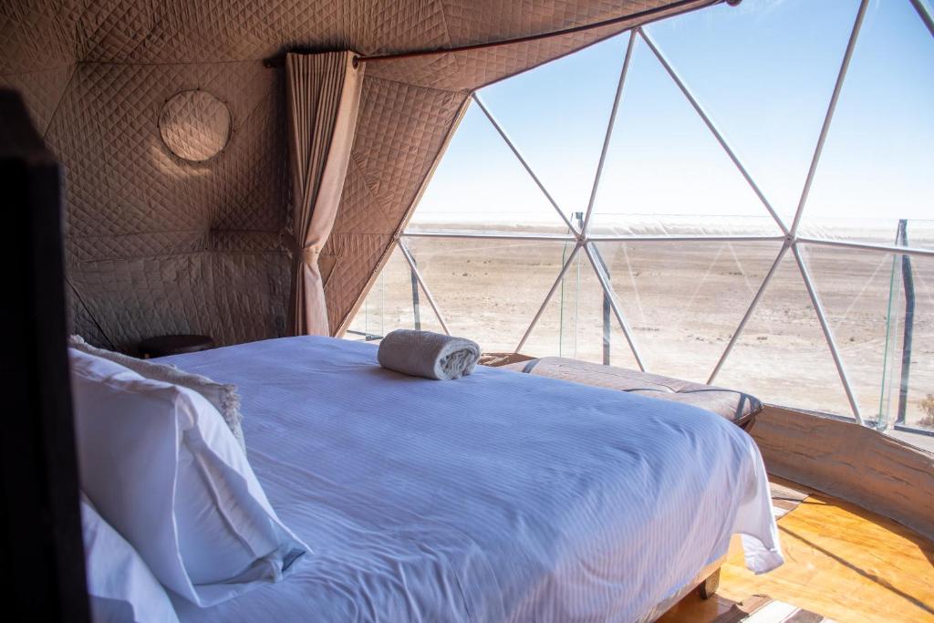 Geodesic-dome bedroom with a white-linen bed, rolled towel on top, and desert view through large glass panels.