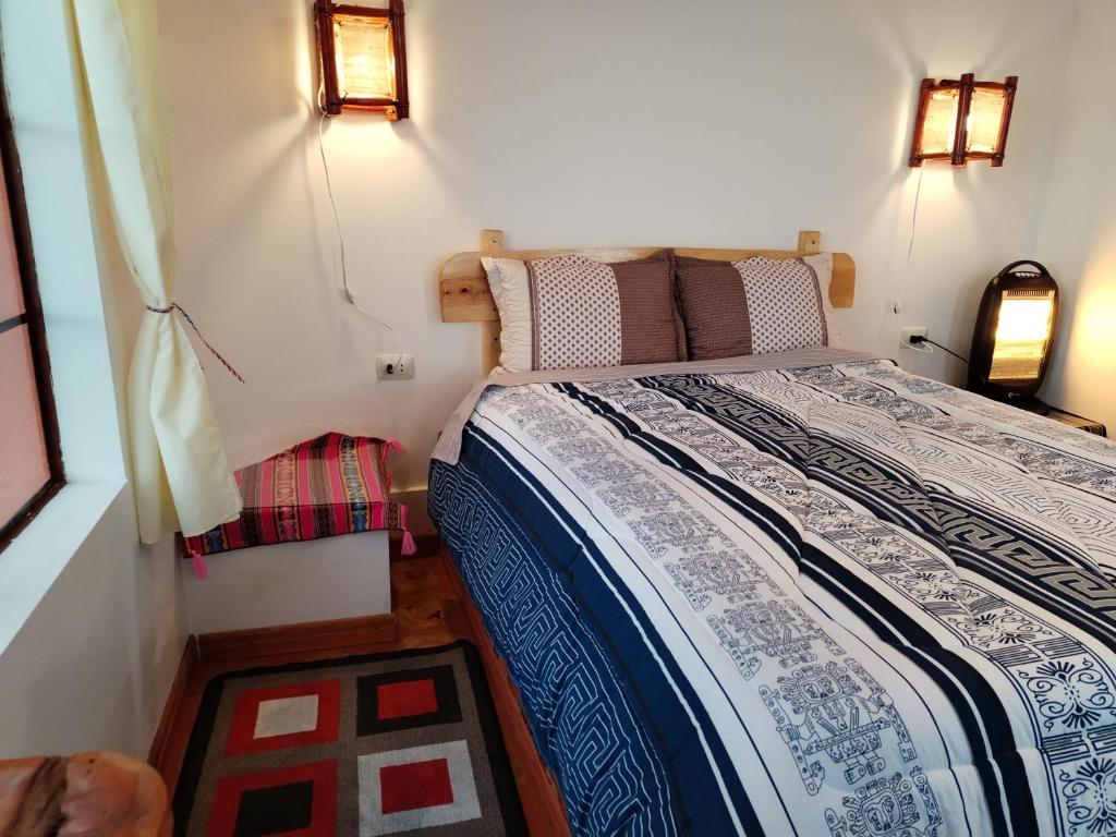 Cozy bedroom with a wooden headboard bed, blue and white patterned quilt, and two beige pillows beneath warm wall lamps. A space heater sits by the right wall. In front is a geometric rug and a window with pale curtains to the left.
