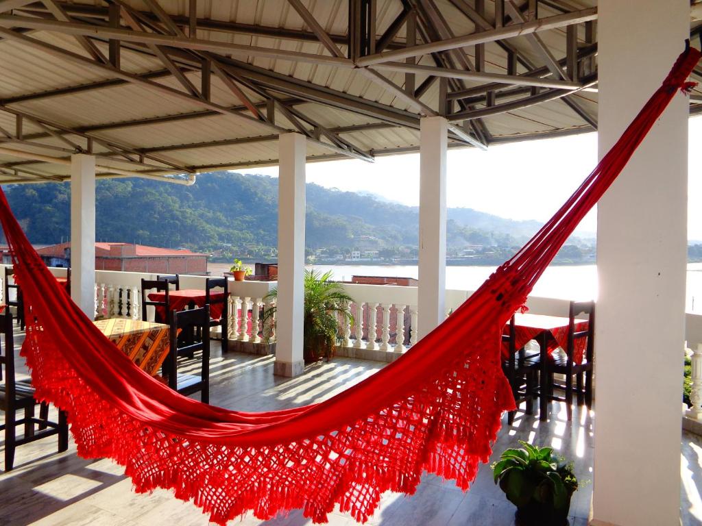 Red woven hammock hangs between white columns on a sunny seaside terrace with tables, chairs, and a railing overlooking water and distant hills.