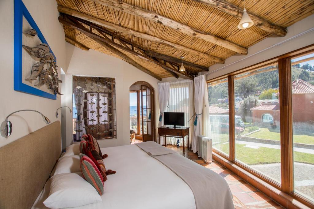 Sunlit hotel bedroom with exposed wooden beams, a large window wall, a king bed with white linens and colorful pillows, and a view of the outdoors.