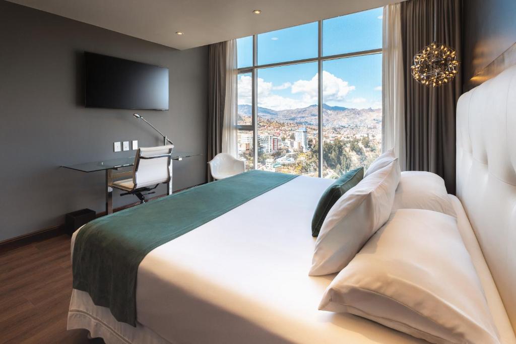 Hotel room with a large white bed, green runner, and pillows, facing a floor-to-ceiling window with a city view and mountains in the distance.