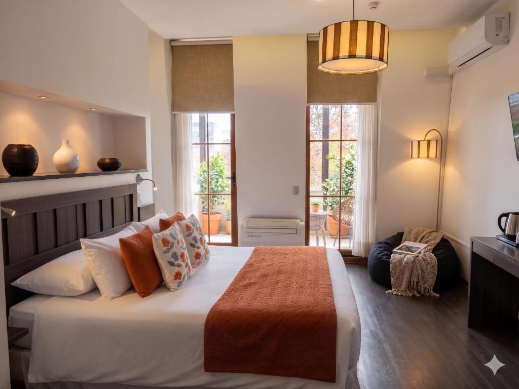 Hotel bedroom with a king bed, orange throw, and patterned pillows, facing glass balcony doors with potted plants outside.