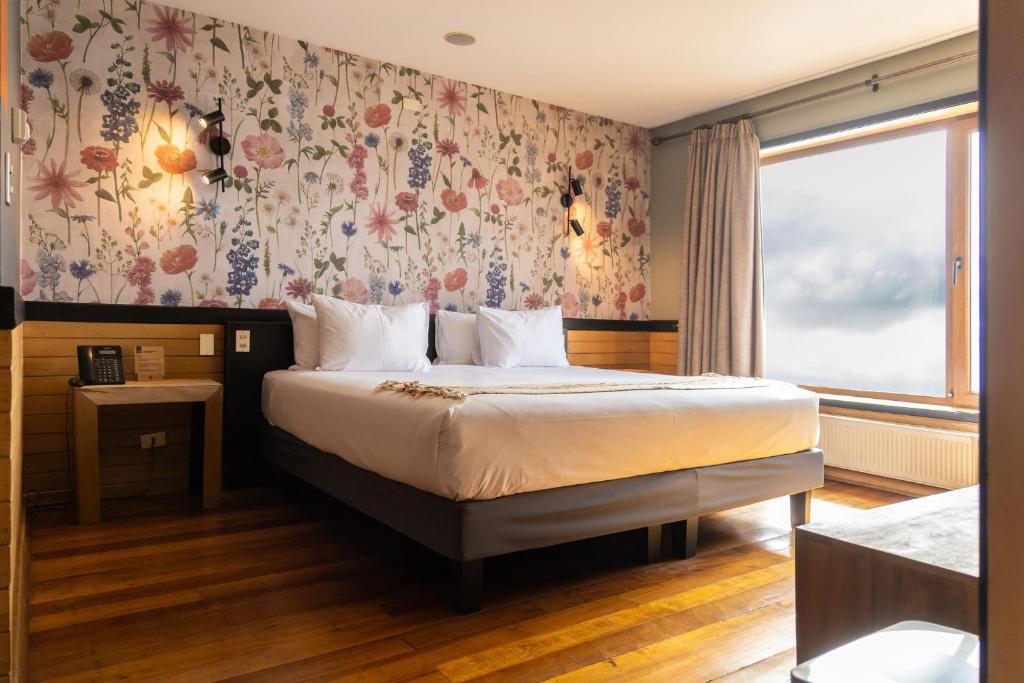 Hotel bedroom with a large bed against a floral wallpaper accent wall, wood nightstand, and a large window with curtains.”