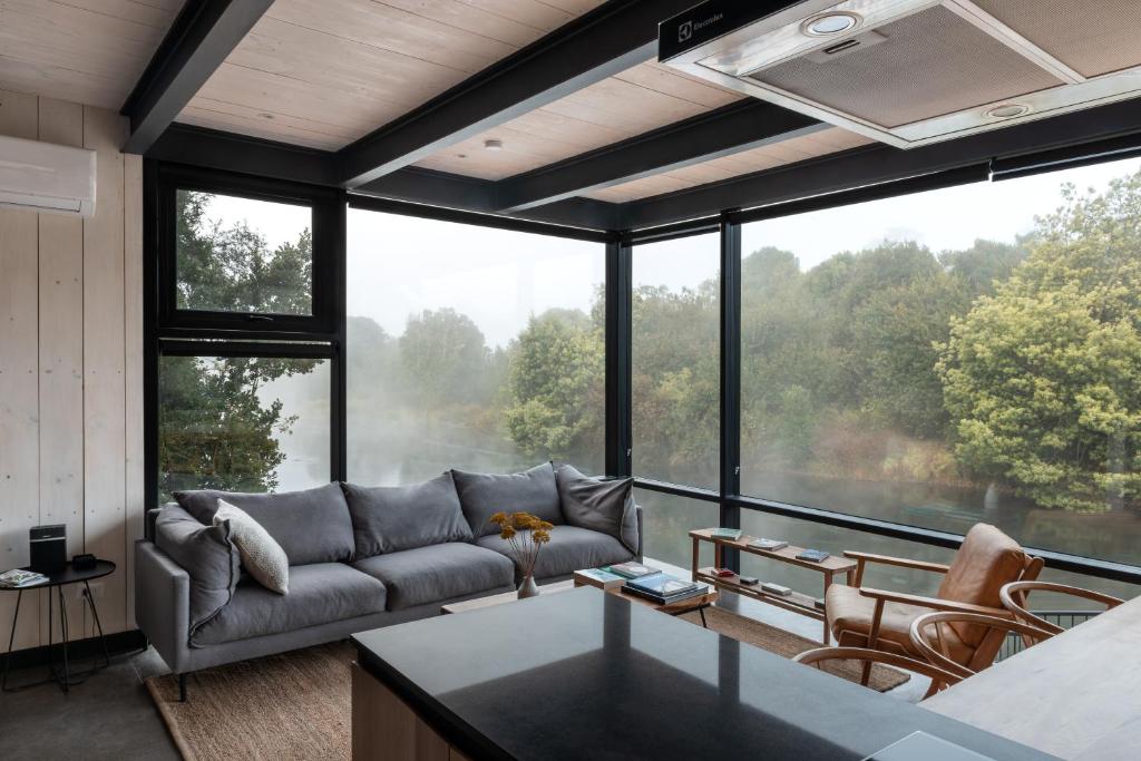 A modern glass-walled living room with a gray sofa facing a lake view and a wooden chair, coffee table, and magazines nearby.