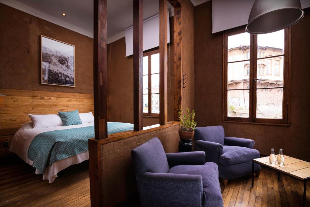 A cozy hotel room with a bed on the left, teal bedspread, and two blue armchairs by a wooden partition and windows on the right.