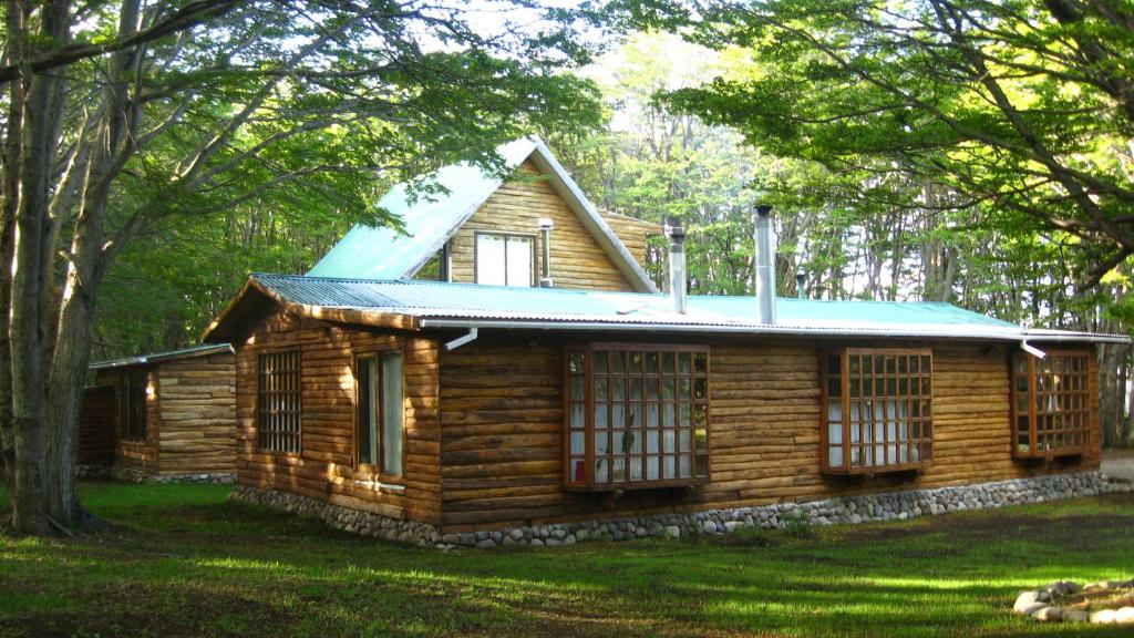 Rustic wooden cabin in a forest, with a green metal roof and large pane windows along a stone foundation.