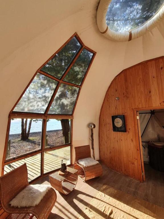 Sunlit interior of a curved A-frame with large angled windows overlooking a beach and trees; two wicker chairs and a small table by the window.