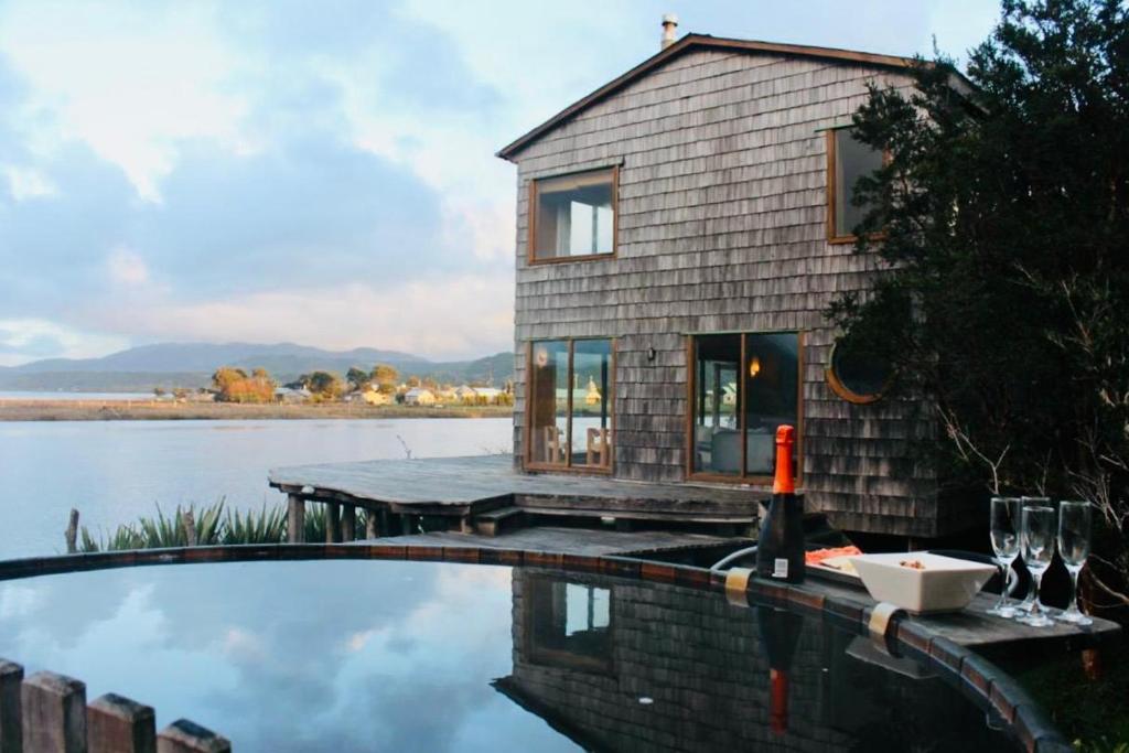 A wooden lakeside house with large windows, seen from a surrounding hot tub with a bottle and glasses on the edge; calm water and hills in the background.